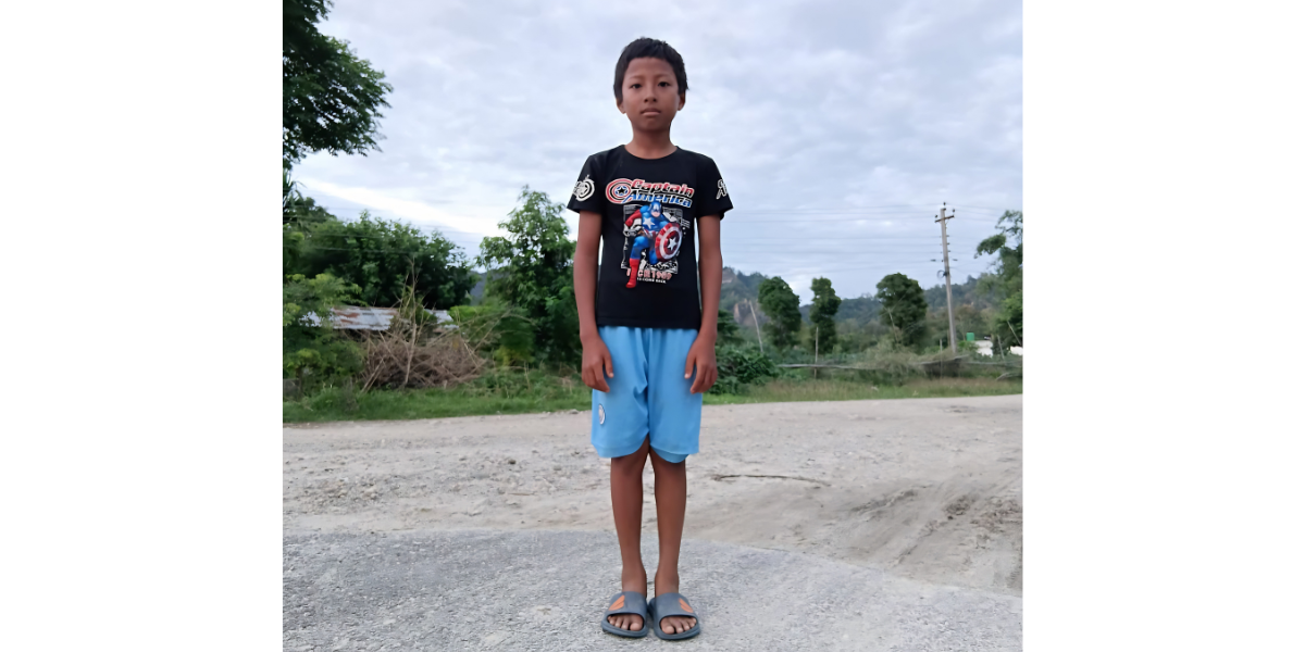 Bhim*, a boy at the center, stands in a road wearing a black Captain America shirt, blue shorts, and slides. Trees and power lines are behind him