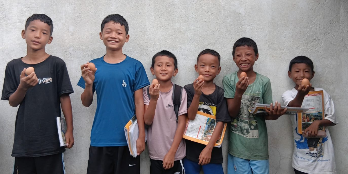Bhim* and his friends, several boys, are lined up against a wall and each are holding an egg in one hand and books in another. They are all smiling