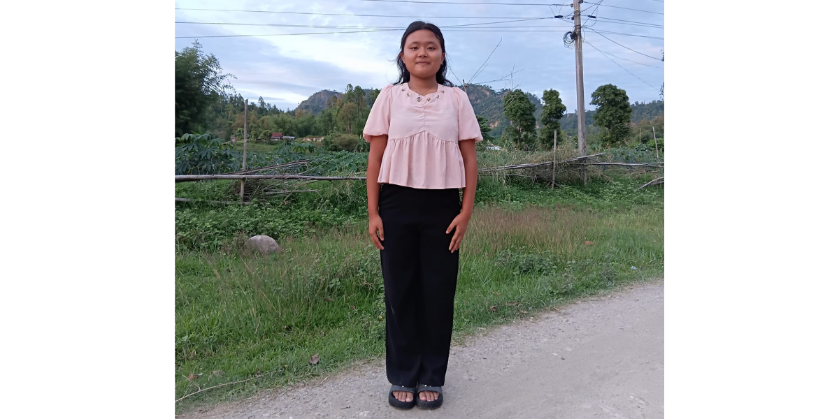 Sabina*, an older girl at the center, stands in a road wearing a pink shirt and black pants. Trees and grass are behind her, along with power lines 